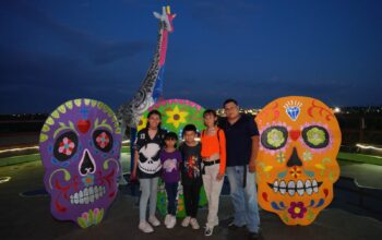 Cinco personas posan en el Zoológico de Nuevo Laredo frente a tres grandes calaveras decorativas de colores y una jirafa ornamental, de noche