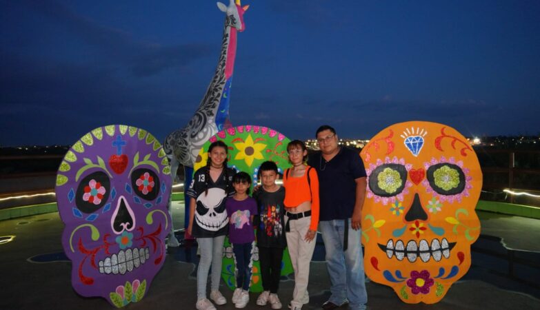 Cinco personas posan en el Zoológico de Nuevo Laredo frente a tres grandes calaveras decorativas de colores y una jirafa ornamental, de noche